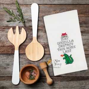 Könnte beinhalten: Ein weißes Küchentuch mit einem Kreuzstichmotiv eines Hauses, einer grünen Godzilla-Figur, die Feuer speit, und dem Text "MAY GODZILLA DESTROY THIS HOME LAST". Dazu eine Bambusgabel, ein Bambuslöffel, eine kleine Holzschale und ein Stößel.