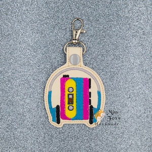 May include: A white leather keychain with a colorful embroidered cassette tape and headphones. The keychain has a silver metal clasp. The text "Miss Foxy Handmade" is embroidered on the bottom.