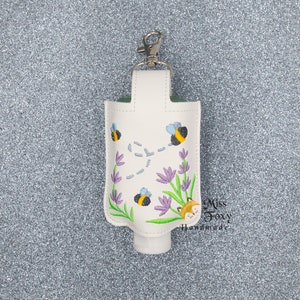 Puede incluir: Soporte blanco para desinfectante de manos con un clip plateado, con bordados de abejas, lavanda y un zorro. El soporte está diseñado para llevar un pequeño frasco de desinfectante. El texto "Miss Foxy Handmade" es visible.