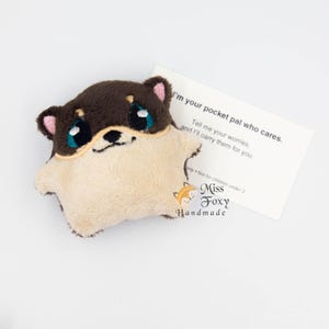 Puede incluir: Un peluche Shiba Inu marrón y crema con grandes ojos azules y orejas rosas. El perro sostiene una pequeña tarjeta que dice: "Soy tu amigo de bolsillo que se preocupa. Dime tus preocupaciones, y las llevaré por ti."