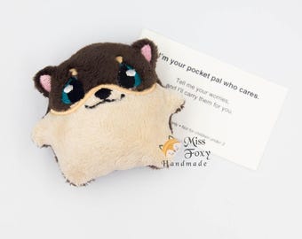 May include: A plush, brown and cream-colored Shiba Inu dog with large blue eyes and pink ears. The dog is holding a small card that reads, "I'm your pocket pal who cares. Tell me your worries, and I'll carry them for you."