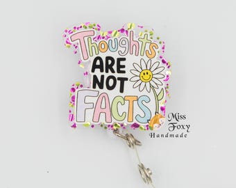 May include: A pink and gold glitter badge reel with a white background and the text "Thoughts Are Not Facts" in colorful letters. A smiling daisy with a yellow center is next to the text. The text "Miss Foxy Handmade" is in the bottom right corner.