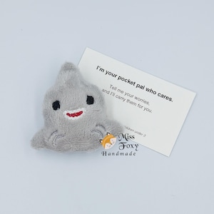 May include: A gray plush shark toy with black eyes and a red smile. A white card next to the shark reads "I'm your pocket pal who cares. Tell me your worries, and I'll carry them for you."