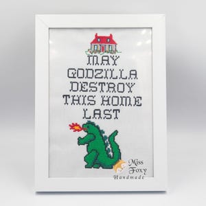 May include: White framed cross-stitch art featuring a green Godzilla breathing fire, a red house, and the text "MAY GODZILLA DESTROY THIS HOME LAST." The artwork is signed "Miss Foxy Handmade."