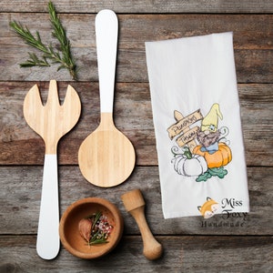 May include: A white kitchen towel with an embroidered design of a gnome holding a sign that says "Pumpkins this way". The gnome is sitting on a pumpkin and there are two other pumpkins in the design. The towel also has the text "Miss Foxy Handmade" at the bottom.