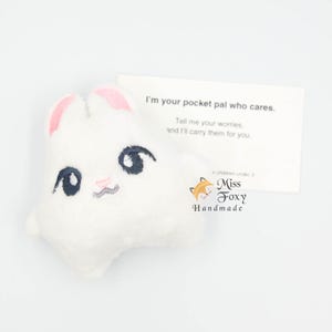 Puede incluir: Un conejito de peluche blanco con el interior de las orejas rosa y rasgos faciales bordados. Una tarjeta junto al conejito dice: "Soy tu amigo de bolsillo que se preocupa. Cuéntame tus preocupaciones y las llevaré por ti."