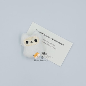 May include: A white plush cat-shaped pocket pal with embroidered facial features. The cat is next to a card that reads "I'm your pocket pal who cares. Give me your worries, I'll carry them for you."