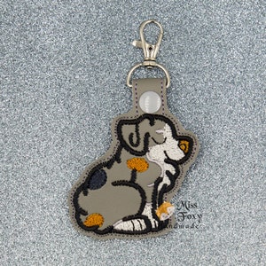 May include: A gray, embroidered dog keychain with black, white, and orange details. The keychain has a silver metal ring and clasp.