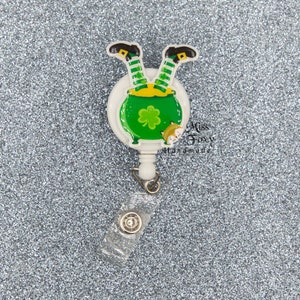 May include: A white retractable badge holder with a green pot of gold design. A cartoon leprechaun's legs are sticking out of the pot. The text "Miss Foxy Handmade" is printed on the holder.