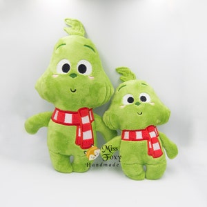 May include: Two plush green Grinch toys with red and white striped scarves. The larger Grinch is standing and the smaller Grinch is sitting. The text "Miss Foxy Handmade" is visible in the bottom right corner of the image.