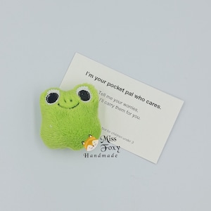 May include: A small, plush green frog with embroidered black eyes and a smiling mouth. A white card next to it reads "I'm your pocket pal who cares. Tell me your worries, I'll carry them for you." A small fox button is also present.