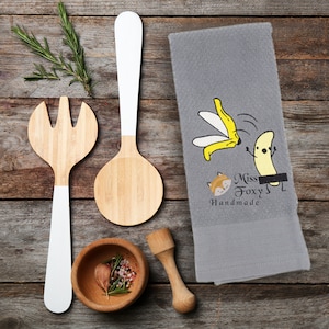 May include: A gray kitchen towel with a cartoon banana slipping on a peel and a fox with the text "Miss Foxy Handmade".