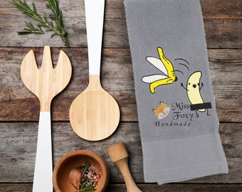 May include: A gray kitchen towel with a cartoon banana slipping on a peel and a fox with the text "Miss Foxy Handmade".