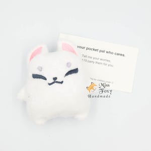 Puede incluir: Un peluche de zorro blanco con el interior de las orejas rosa y rasgos faciales bordados en negro. Una tarjeta al lado dice "your pocket pal who cares" y "Miss Foxy Handmade".