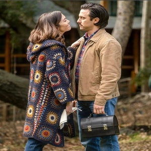 May include: A couple standing close together, the woman is wearing a long, crocheted cardigan with a colorful pattern, the man is wearing a brown jacket and blue jeans, he is holding a black metal lunchbox.