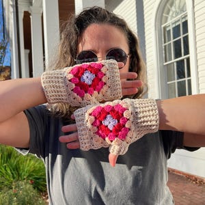Puede incluir: Un par de guantes sin dedos de color beige con cuadrados de crochet rosa, rojo y morado. Los guantes están hechos con un hilo texturizado y tienen un diseño sencillo.