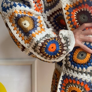 May include: A white crochet cardigan with a colorful geometric pattern. The cardigan features a variety of colors, including orange, brown, blue, and green. The cardigan is made of a soft, lightweight yarn.