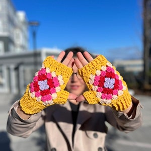 Puede incluir: Un par de guantes sin dedos de color amarillo hechos a crochet con detalles en rosa, rojo y morado. Los guantes tienen un patrón de granny square.