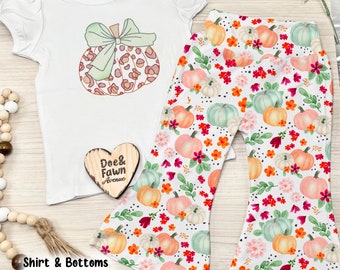 May include: White short-sleeve shirt with a pumpkin design and bell-bottom pants. The shirt features a pumpkin with a green bow. The pants have a white background with a pumpkin and floral pattern. The text "Dee & Fawn Avenue" is on a wooden heart.
