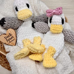 May include: Two handmade crochet plush ducks with white bodies, yellow beaks and feet, and grey wings. One duck features a pink crochet bow on its head. A heart-shaped wooden plaque with the text Dee & Fawn Avenue is placed next to the plush toys.