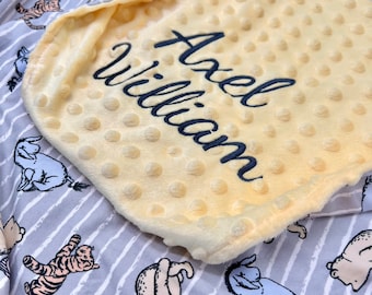 May include: A yellow and grey baby blanket featuring a Winnie the Pooh design. The blanket has a soft, plush texture and is personalised with the name "Axel William".