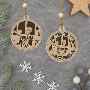 Puede incluir: Dos adornos navideños de madera con temática de ballet. Cada uno presenta la silueta de un bailarín y un nombre: Viviana y Leonard. Los adornos redondos tienen un acabado marrón claro y están suspendidos por una cuerda. También hay copos de nieve decorativos y una estrella de madera.