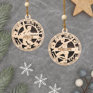 May include: Two wooden Christmas ornaments with a dancer silhouette and the names "Jerico" and "Penelope". The ornaments are round with a dancer design and text around the edge. They hang from a string with a wooden bead.