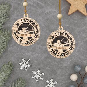 Peut inclure: Deux décorations de Noël en bois sur le thème du tir à l'arc, chacune avec un nom gravé. Les ornements circulaires présentent une silhouette d'archer et des termes de tir à l'arc. Ils sont suspendus à une ficelle avec une perle en bois.