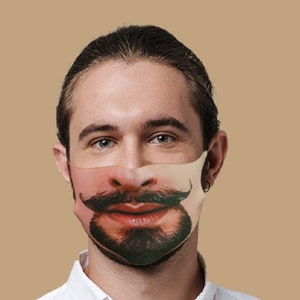 May include: A man wearing a white button-down shirt and blue pants is wearing a face mask with a printed image of a man with a mustache and beard.