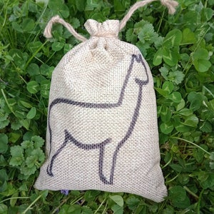 May include: A small burlap bag with a drawstring closure, featuring a black silhouette of an alpaca. The bag is sitting on a bed of green clover.