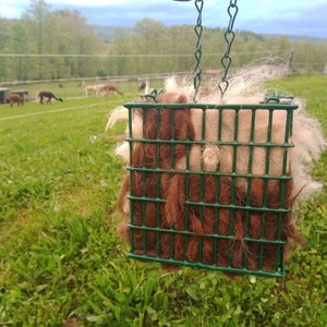 Pode incluir: Uma gaiola de arame verde cheia de l&atilde; de alpaca marrom e branca pendurada em uma corrente. A gaiola est&aacute; em um campo com grama verde e uma cerca no fundo.