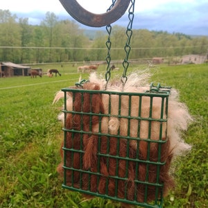 Pode incluir: Uma gaiola de arame verde cheia de l&atilde; de alpaca marrom e branca pendurada em um gancho de metal. A gaiola &eacute; suspensa por uma corrente.