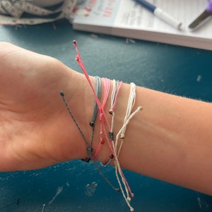 Op de afbeelding: Een close-up van een pols met verschillende kleurrijke vriendschapsarmbanden met kleine kralen. De armbanden zijn gemaakt van dun touw en zijn in een knoop geknoopt.