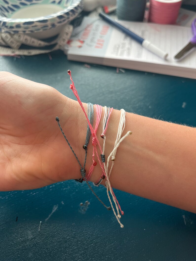 Op de afbeelding: Een close-up van een pols met verschillende kleurrijke vriendschapsarmbanden met kleine kralen. De armbanden zijn gemaakt van dun touw en zijn in een knoop geknoopt.