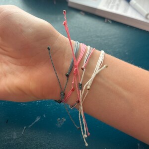 Op de afbeelding: Een stapel kleurrijke vriendschapsarmbanden met zwarte en gouden kralen. De armbanden zijn gemaakt van dun touw en zijn om een pols gebonden.