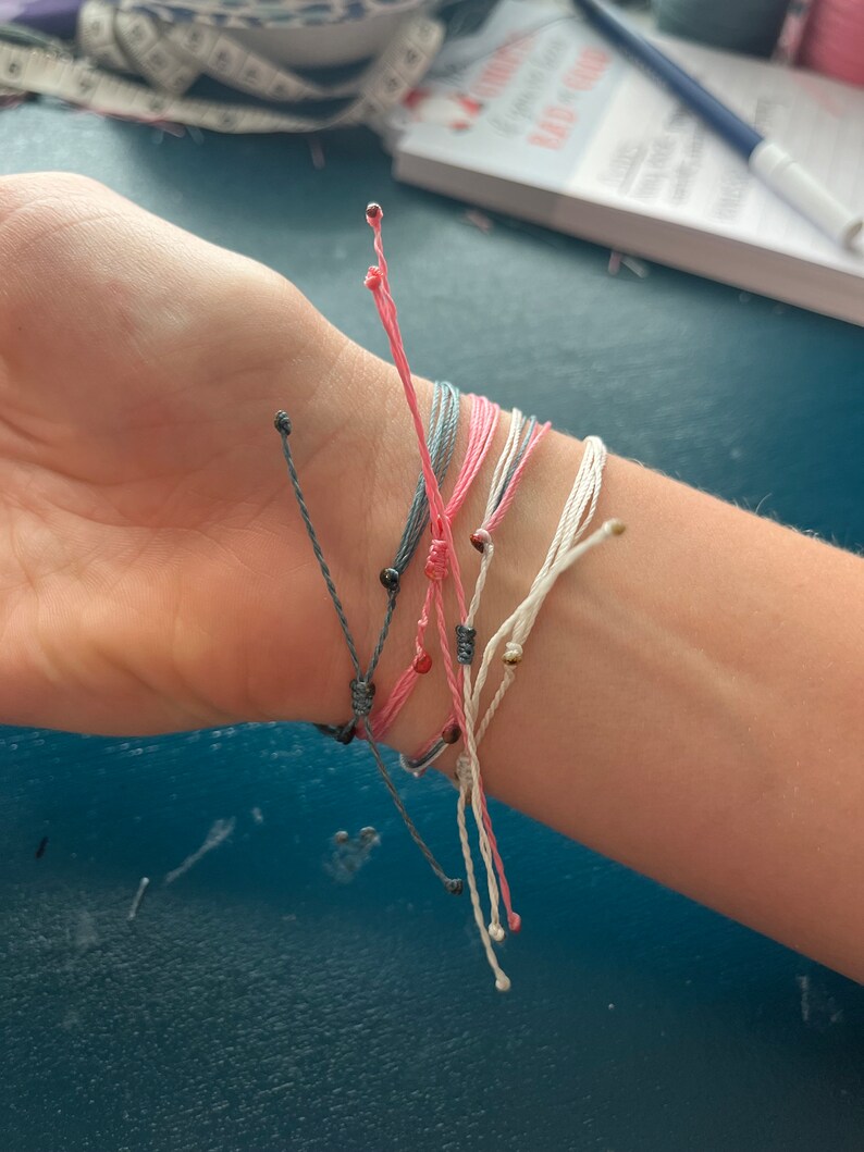 Op de afbeelding: Een stapel kleurrijke vriendschapsarmbanden met zwarte en gouden kralen. De armbanden zijn gemaakt van dun touw en zijn om een pols gebonden.