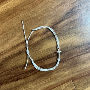 Op de afbeelding: Witte armband van touw met een zilveren kruisbedel.