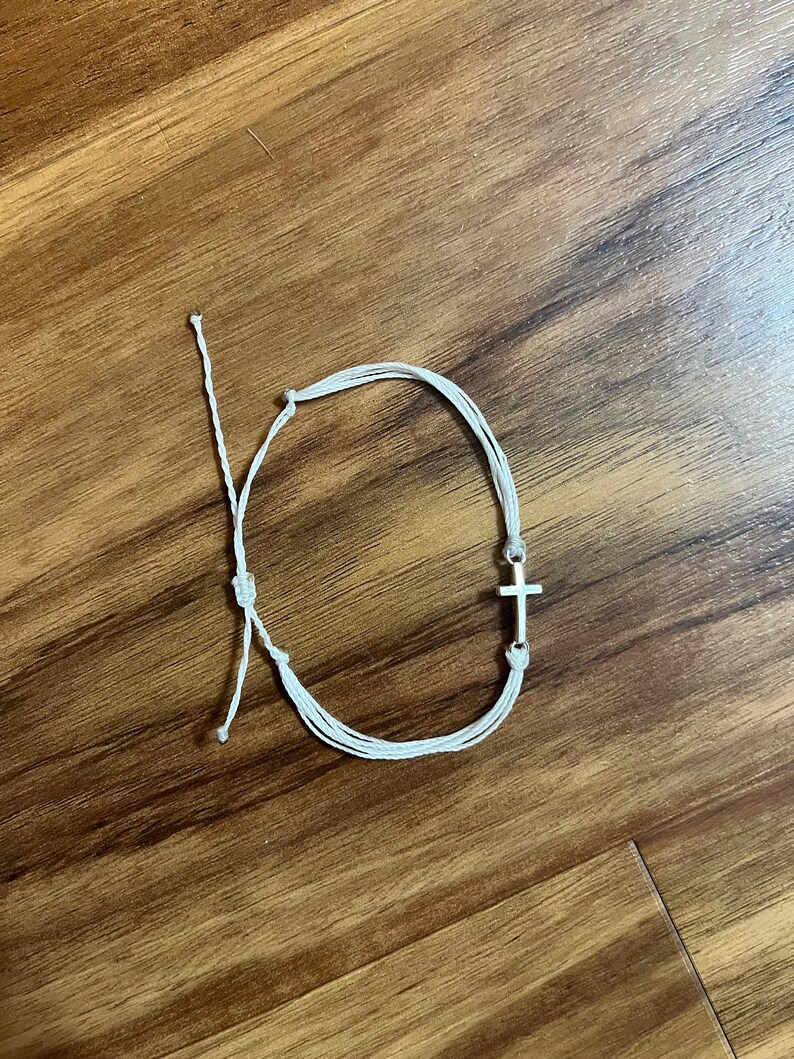 Op de afbeelding: Witte armband van touw met een zilveren kruisbedel.