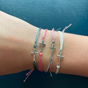 Op de afbeelding: Vier vriendschapsarmbanden met zilveren kruisbedels. De armbanden zijn in tinten roze, grijs en wit.