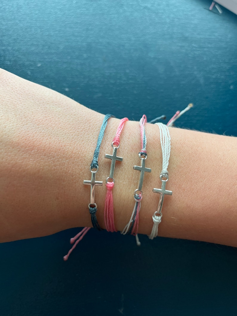 Op de afbeelding: Vier vriendschapsarmbanden met zilveren kruisbedels. De armbanden zijn in tinten roze, grijs en wit.