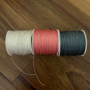 Op de afbeelding: Drie spoelen garen, een witte, een roze en een donkergrijze.