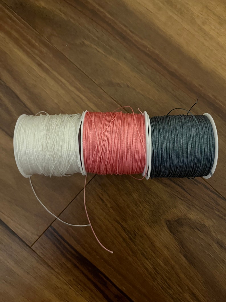 Op de afbeelding: Drie spoelen garen, een witte, een roze en een donkergrijze.