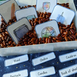May include: A white plastic container filled with brown beans, with illustrated cards depicting magical symbols and objects such as a wand, chalice, and cauldron. Additional labels with text are scattered around the container, including "Candles" and "Book of shadows".