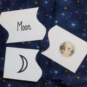 May include: Three white cards with black outlines and rounded edges. The top card has the word "Moon" written in black. The middle card has a black and white illustration of a crescent moon. The bottom card has a black and white illustration of a full moon.