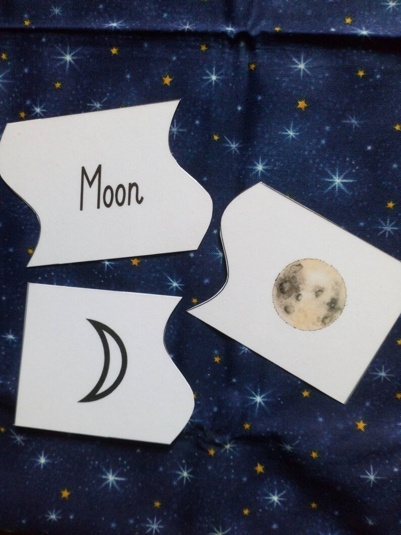 May include: Three white cards with black outlines and rounded edges. The top card has the word "Moon" written in black. The middle card has a black and white illustration of a crescent moon. The bottom card has a black and white illustration of a full moon.
