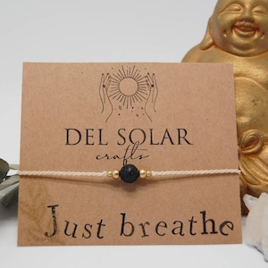Könnte beinhalten: Ein weißes Armband aus Schnur mit einer schwarzen Lavasteinperle und goldenen Perlen. Das Armband liegt auf einer braunen Karte mit dem Text "DEL SOLAR crafts" und "Just breathe".