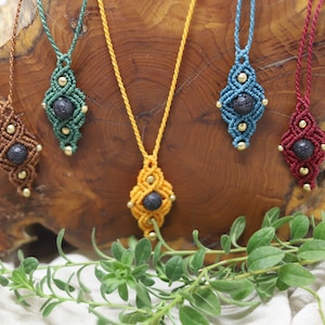 Könnte beinhalten: Fünf Makramee-Halsketten mit Lava-Stein-Anhängern. Die Halsketten sind in verschiedenen Farben: Braun, Grün, Gelb, Blau und Rot. Die Anhänger sind alle gleich groß und in der gleichen Form, und sie sind aus dunkelgrauem Lavastein.