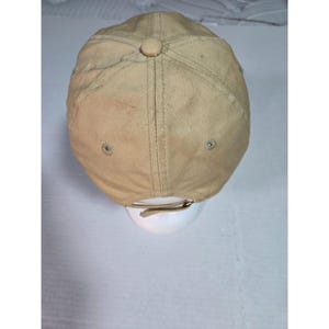 May include: A tan baseball cap featuring a six-panel construction with stitched seams, a fabric-covered button at the crown, and metal eyelets for ventilation. The back includes an adjustable strap with a metal buckle closure for a secure fit.