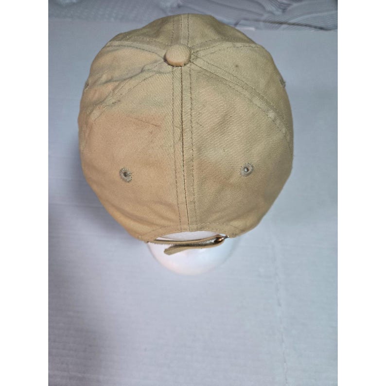 May include: A tan baseball cap featuring a six-panel construction with stitched seams, a fabric-covered button at the crown, and metal eyelets for ventilation. The back includes an adjustable strap with a metal buckle closure for a secure fit.