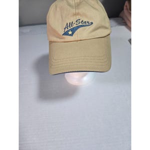 May include: A tan baseball cap featuring the embroidered text All-Star in dark blue script. The design includes a dark blue swoosh shape underneath the text with a small yellow star centered within it. The cap has a curved brim and stitching.